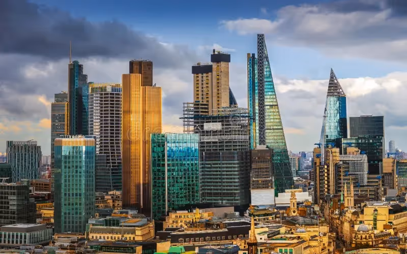 View of modern skyscrapers in a city skyline under a partly cloudy sky with warm sunlight reflecting off the buildings.