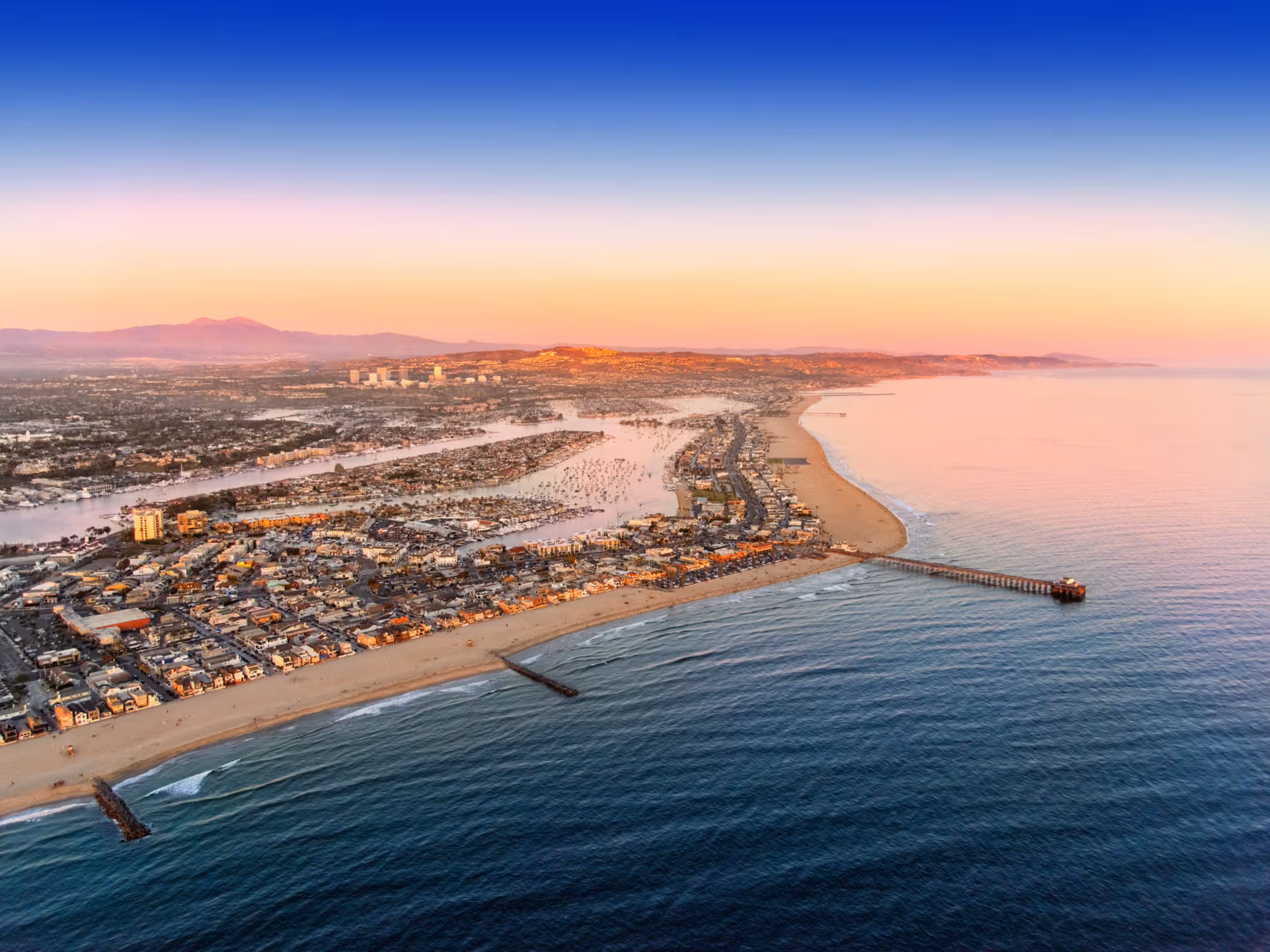 Aerial view of a coastal city at sunset with sandy beaches, a pier extending into the ocean, and residential areas by the water.