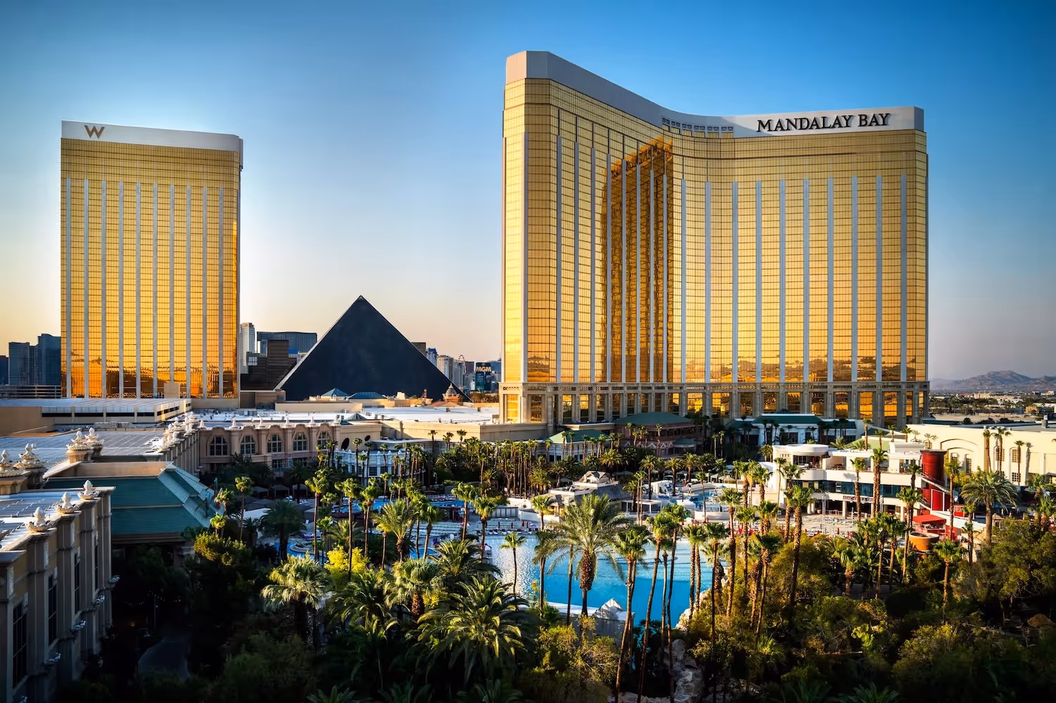 Mandalay Bay hotel and casino towers in Las Vegas with a large outdoor pool and palm trees in the foreground under a clear blue sky.