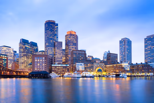 Boston skyline at dusk with illuminated buildings reflected on the water.