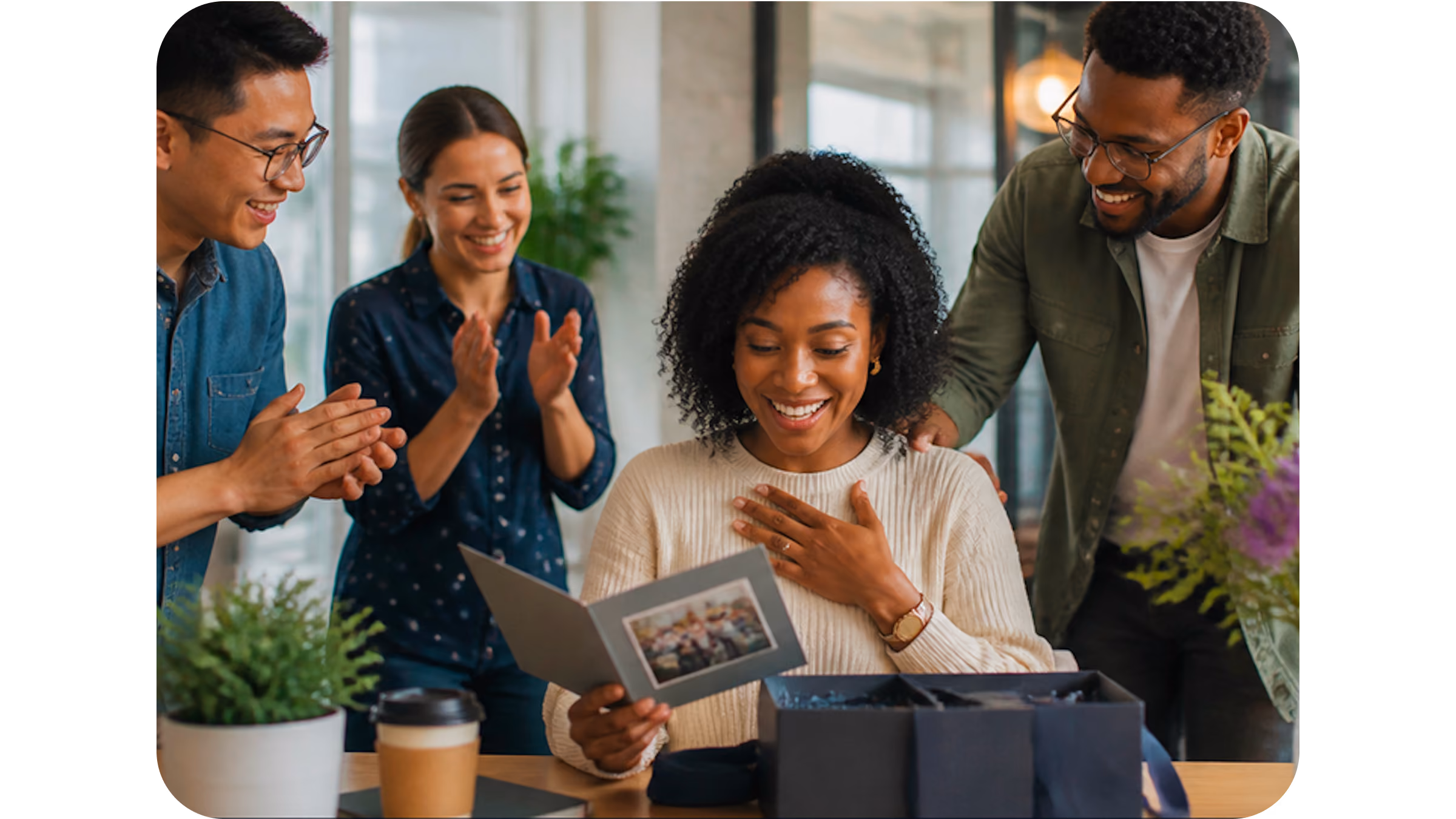 Smiling woman receives a card and gift box while coworkers applaud and congratulate her indoors.
