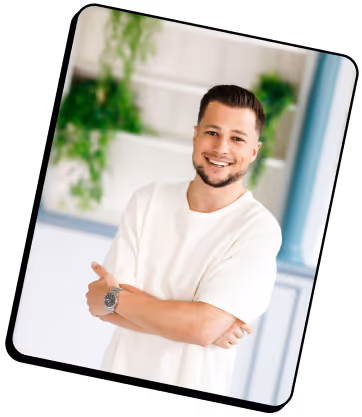 Homme souriant, les bras croisés, vêtu d'un t-shirt blanc et portant une montre, debout à l'intérieur avec des plantes vertes en arrière-plan.