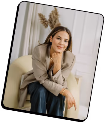 Femme aux longs cheveux bruns, vêtue d'un blazer beige et d'un pantalon foncé, assise dans un fauteuil crème, le menton posé sur la main, souriant doucement.