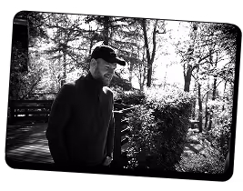 Homme portant une casquette et des vêtements sombres, debout à l'extérieur près d'une balustrade en bois, avec des arbres et des buissons en arrière-plan, en noir et blanc.