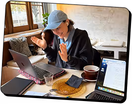 Femme portant une casquette, souriante et gesticulant, assise à une table avec des ordinateurs portables, du café et des collations.