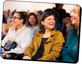 Three women sitting closely together smiling and engaging in conversation at an indoor event.