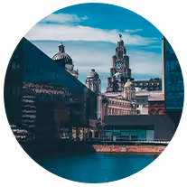 View of historic domed and clock towers reflected in modern glass buildings by a waterfront.