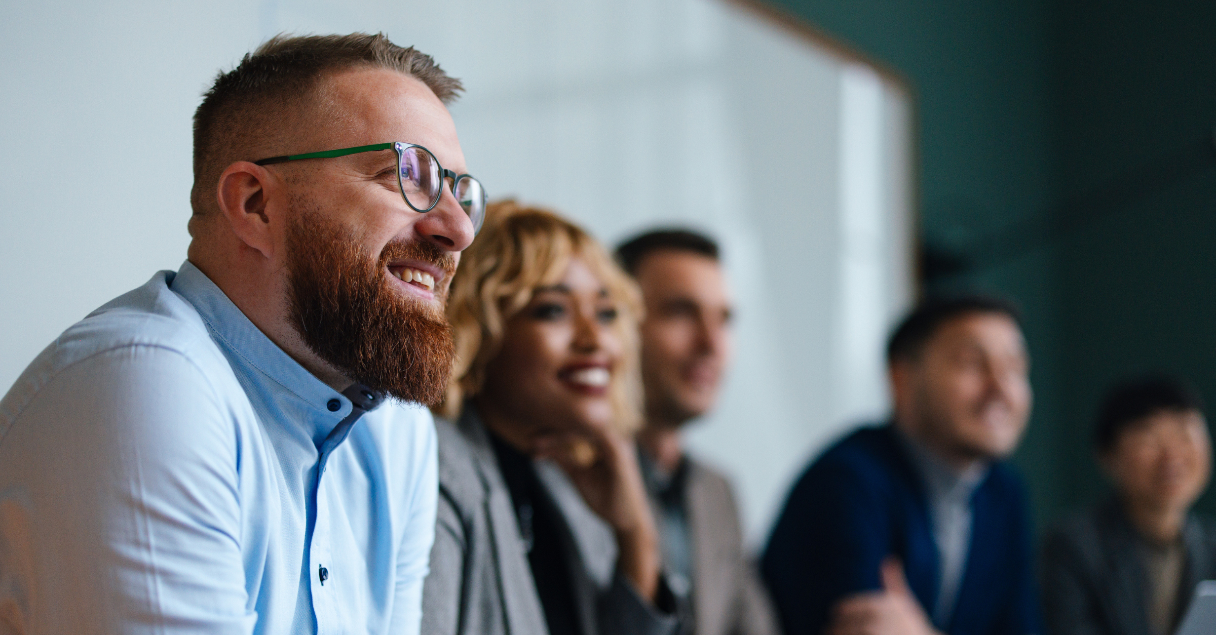 A diverse group of five professionals sitting in a row, smiling and engaged, with a bearded man wearing green-framed glasses in the foreground.
