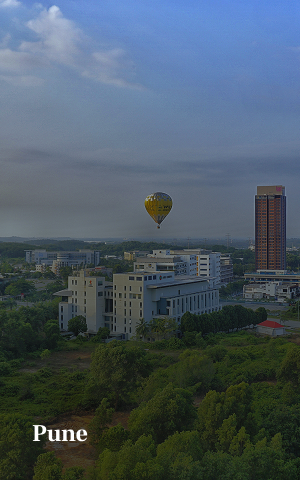 A yellow hot air balloon floating above buildings and greenery in Pune city under a partly cloudy sky.