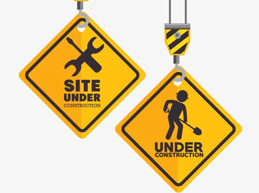 Deux panneaux jaunes en forme de losange suspendus, un avec une clé et un tournevis croisés et le texte 'SITE UNDER CONSTRUCTION', l'autre avec une silhouette de travailleur avec une pelle et le texte 'UNDER CONSTRUCTION'.