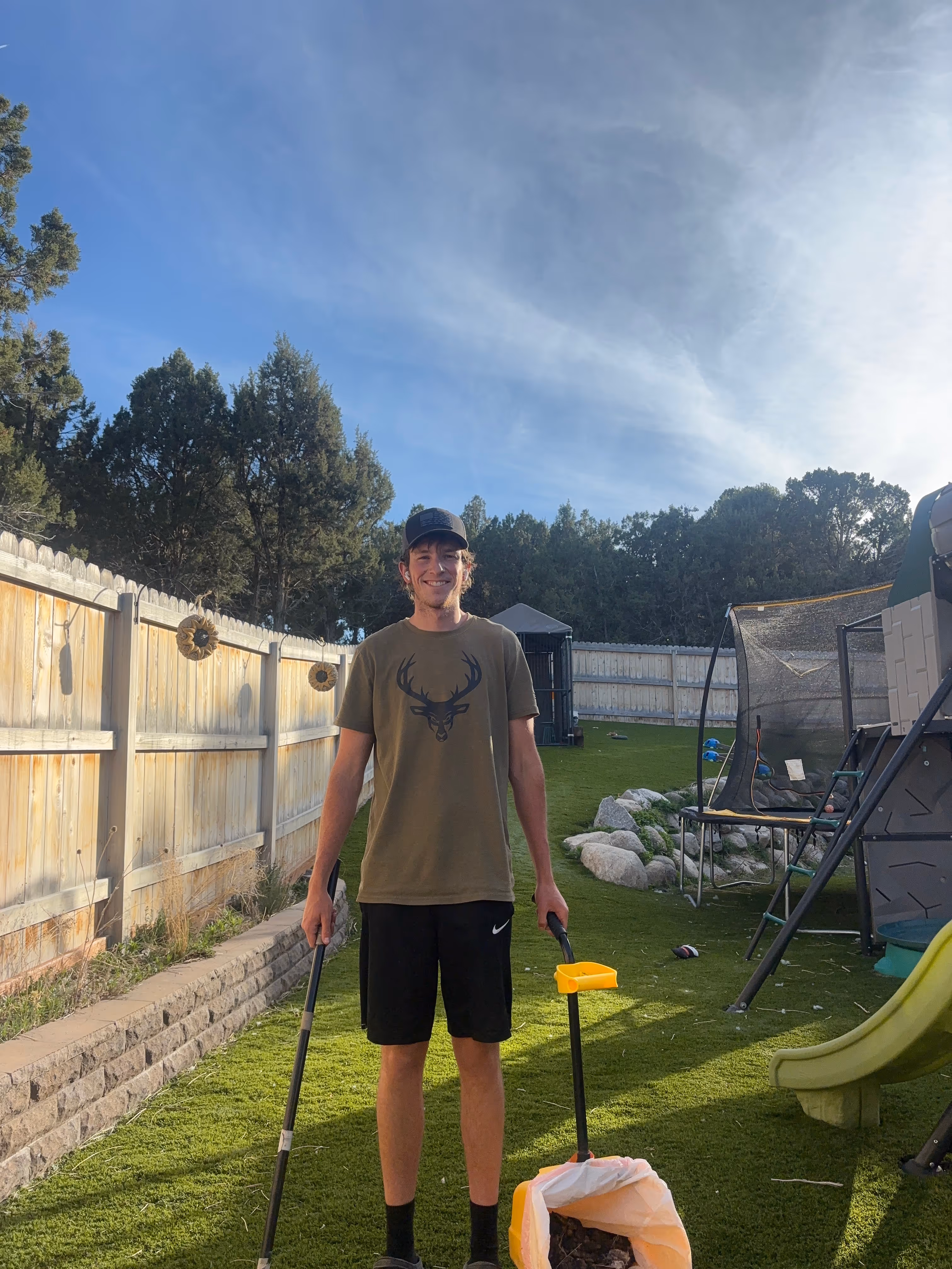 Spencer Scoops technician standing on artificial turf in a residential backyard in Herriman, Utah, holding dog waste removal tools and smiling, with a collection bag at his feet