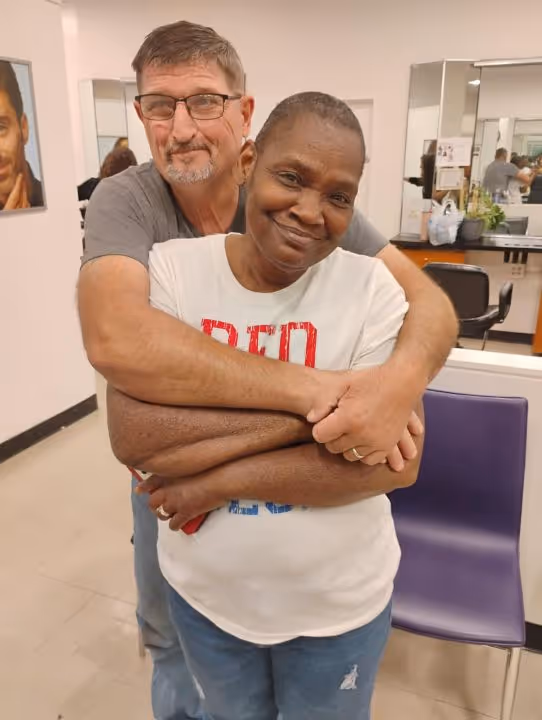 Smiling couple embracing indoors, with the man hugging the woman from behind in a room with salon-style mirrors and chairs.