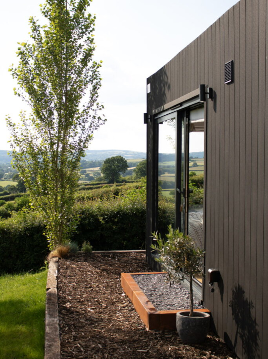 Exterior side view of a modern black cladded container conversion with a glass door, potted plant, and wooden steps, set in a landscaped garden overlooking a scenic countryside landscape with rolling hills and trees.