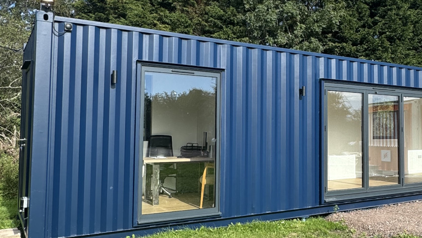 Exterior of a modern blue converted shipping container with large glass windows and doors, situated on grass and gravel near tall evergreen trees, with visible interior office and living space.