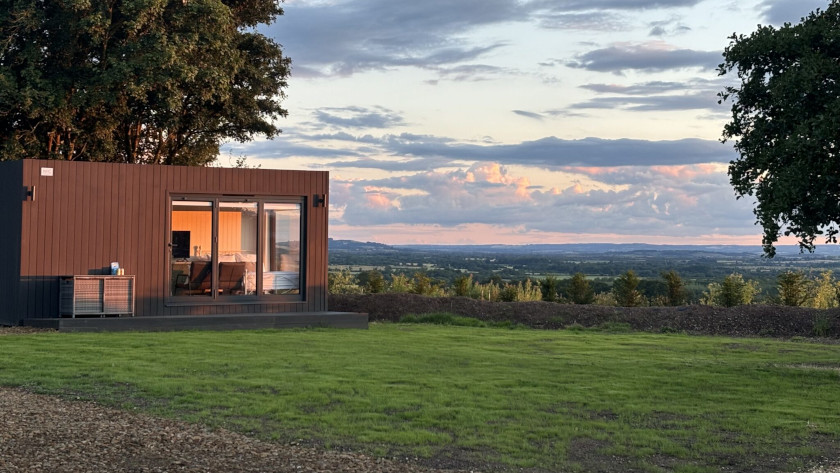 Modern container home with large glass doors and a wooden exterior, situated on a green lawn with expansive views of the countryside under a colorful sunset sky