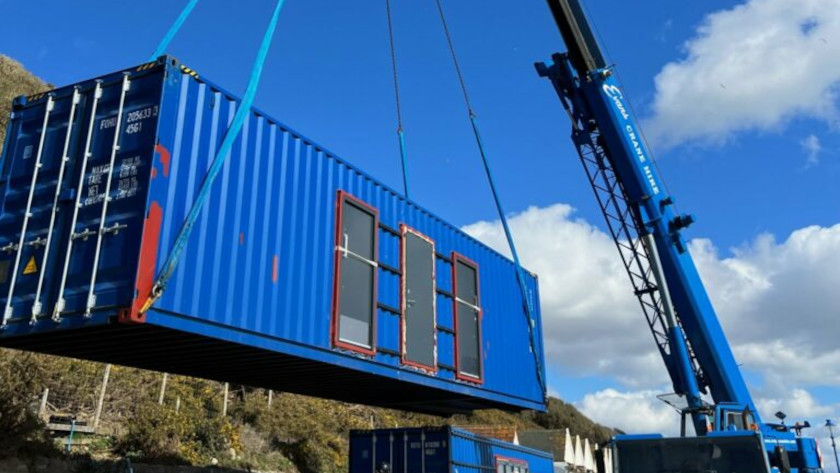 A blue crane lifts a bright blue converted shipping container with window and door openings into place at a construction site, under a sunny sky with scattered clouds.