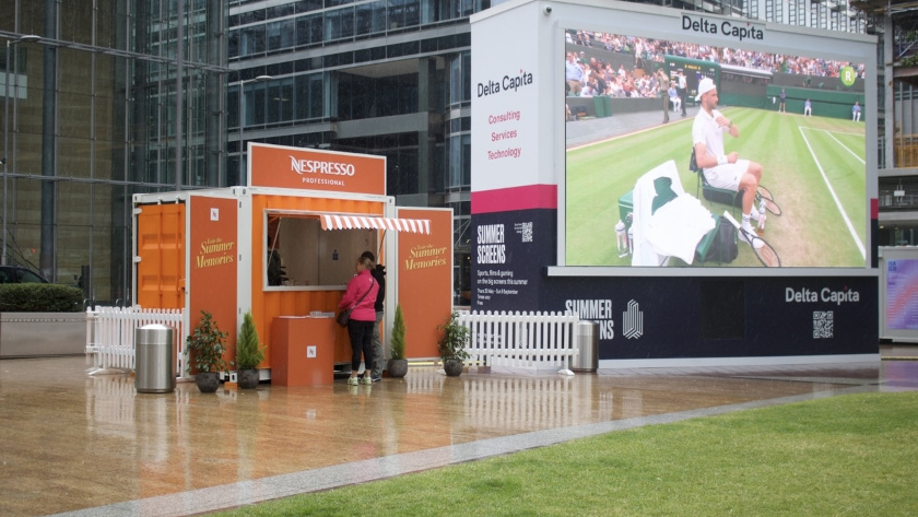 right orange Nespresso promotional kiosk made from a converted shipping container, labeled “Summer Memories,” with a striped canopy and potted plants, set up in an urban plaza beside a large outdoor screen showing a live tennis match.