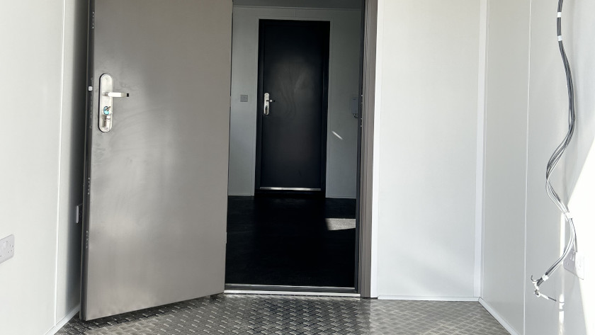 Interior of a secure converted shipping container unit featuring a heavy-duty metal door, diamond plate flooring, white walls, and visible electrical conduit, with a second closed security door visible in the adjacent room.
