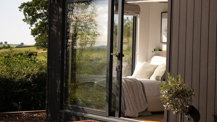 Modern container accommodation unit with large glass doors, showing a cozy interior bedroom with natural light, soft furnishings, and countryside views.