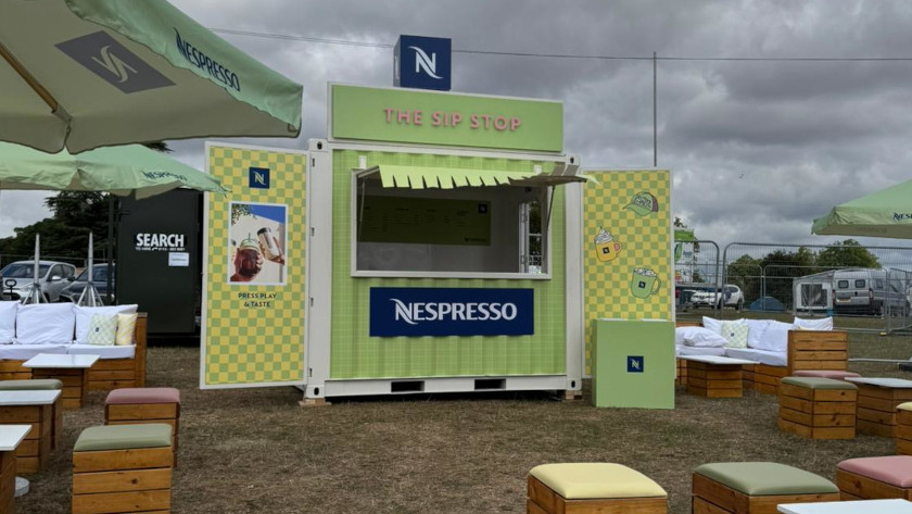 Outdoor Nespresso pop-up café named "The Sip Stop" housed in a converted shipping container painted in green and yellow checkered patterns, with branding and graphics. Surrounded by cushioned wooden seating, parasols, and event fencing under a cloudy sky. 