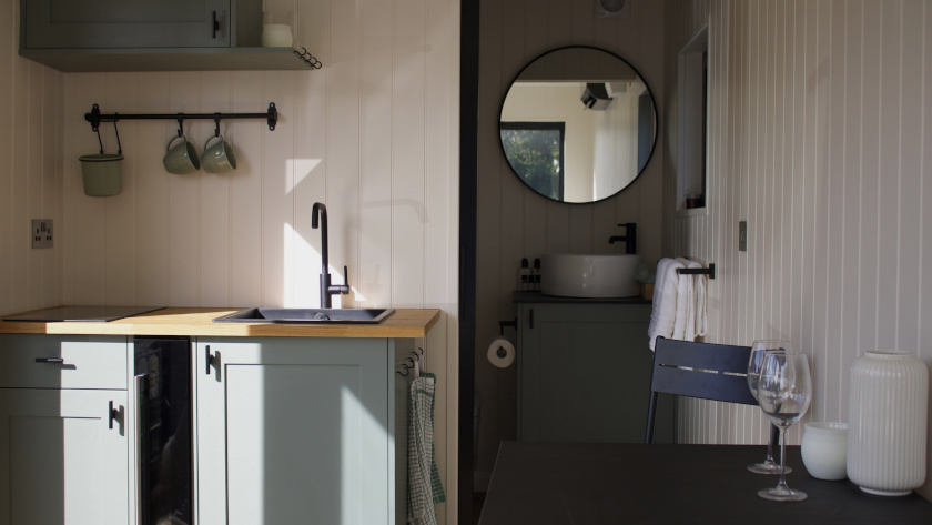 Compact modern kitchen and bathroom area in a container conversion, featuring sage green cabinets, wooden countertop, black sink and tap, wall-mounted mugs, and a bathroom with a round mirror, basin sink, and towel rail in the background.