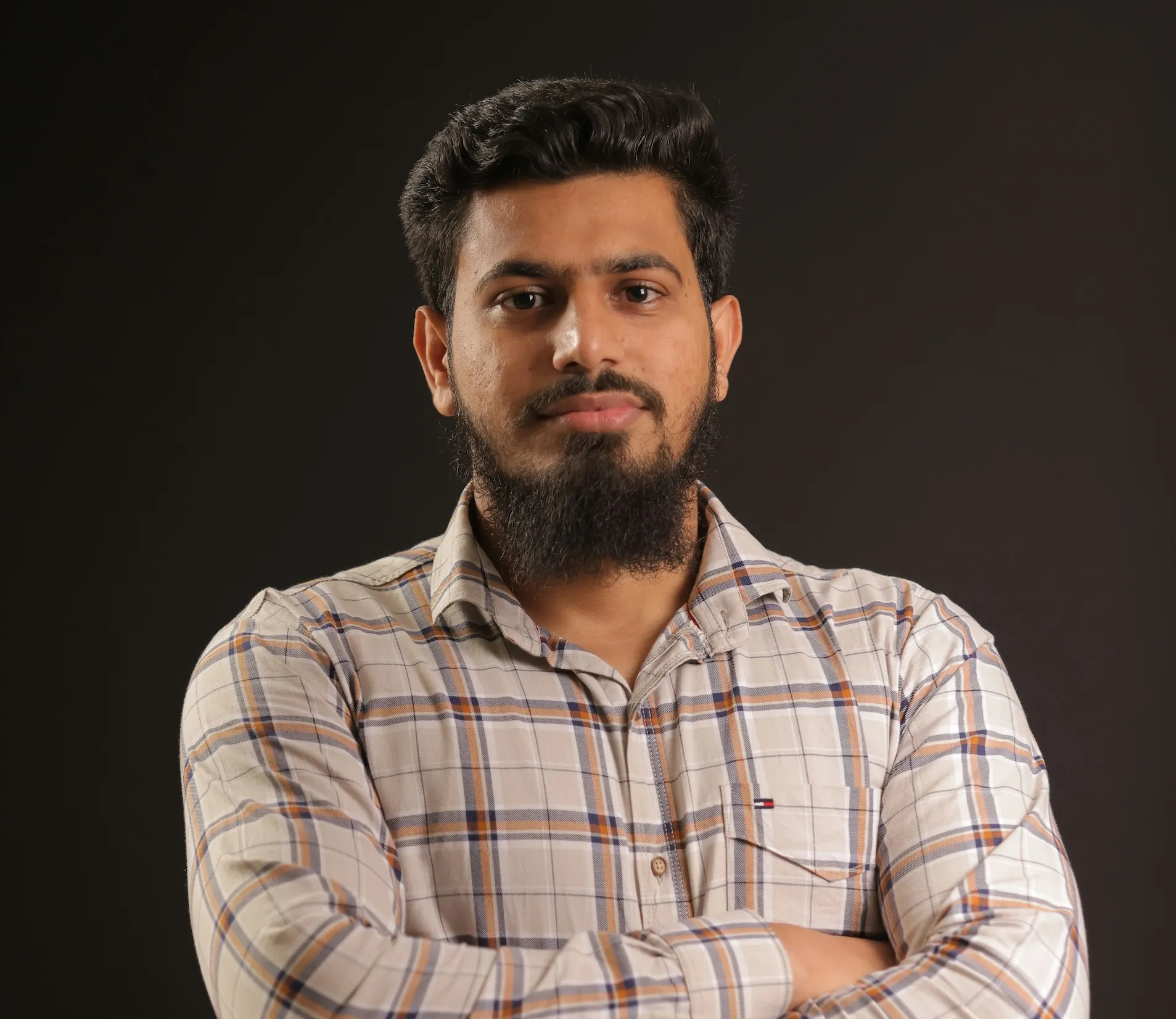 Person with beard in plaid shirt posing with crossed arms against dark background
