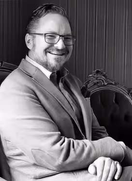 Leende man i kostym sitter vid en dekorativ stol i svartvitt foto