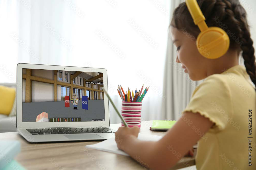 Young girl wearing yellow headphones and a yellow shirt sitting at a desk, writing in a notebook while looking at a laptop screen showing a Minecraft Education game.