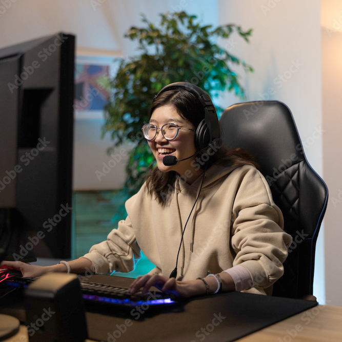 Smiling young woman wearing glasses and headset, playing video games on a computer in a gaming chair.