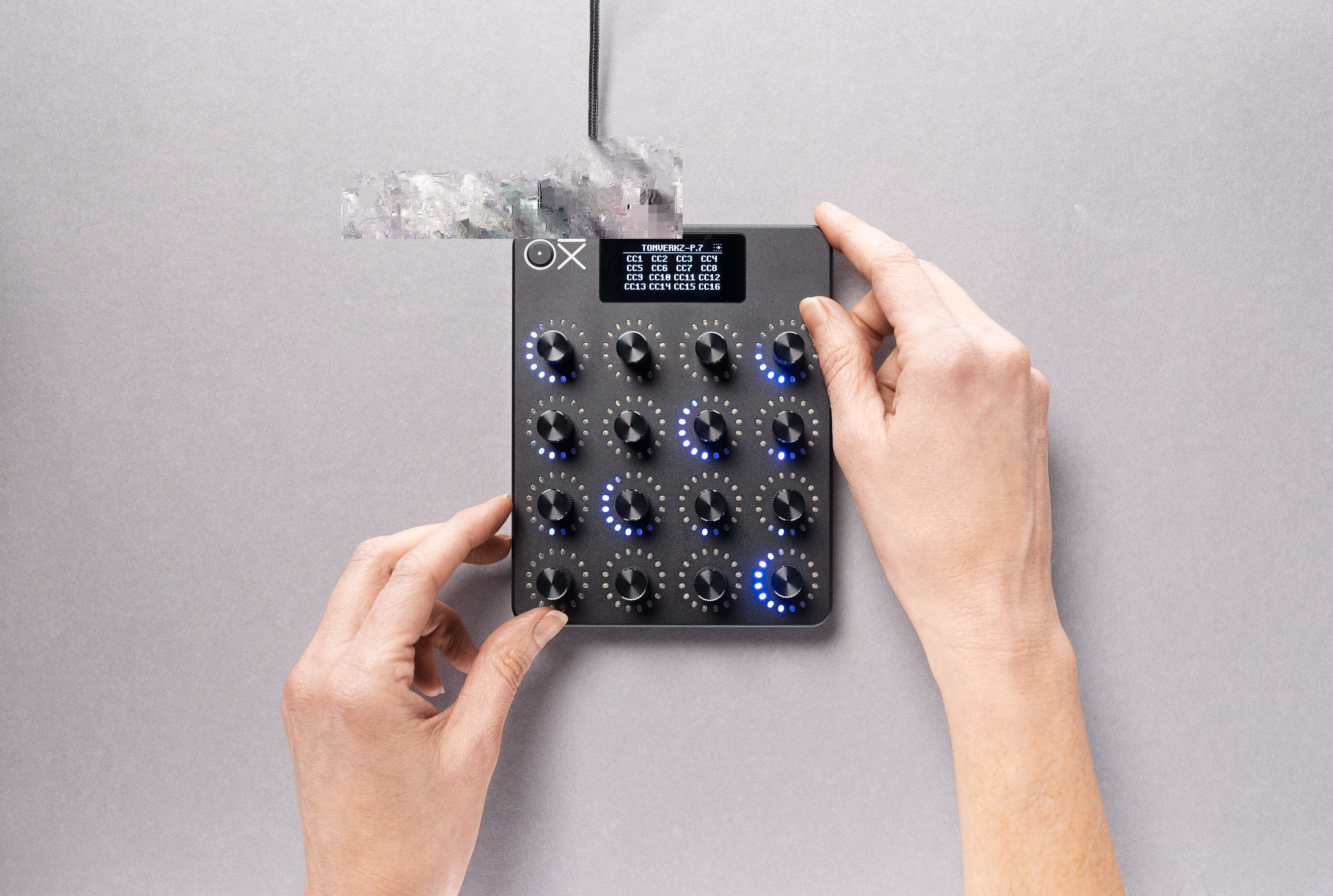 Hands adjusting knobs on a compact black audio or MIDI controller with a small display showing text and blue LED indicators around some knobs.