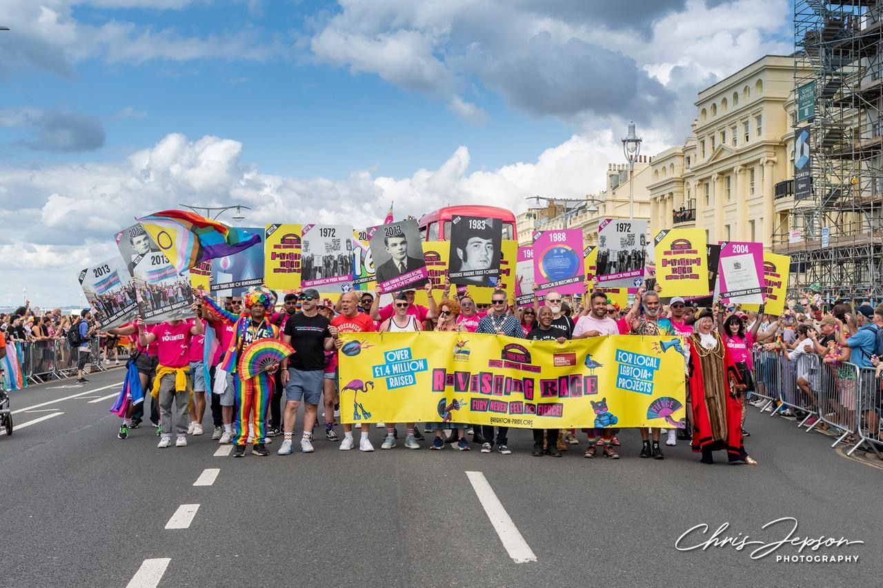 Pride Community Foundation's walking group, a large group of people, march in a Pride parade on a wide street lined with cheering crowds. They hold colourful signs highlighting key LGBTQ+ history dates and achievements, and a bright yellow banner that reads “Ravishing Rage – Over £1.4 Million Raised – For Local LGBTQ+ Projects.” Participants wear vibrant outfits, including rainbow clothing and costumes, while waving Pride flags. The atmosphere is celebratory and empowering under a partly cloudy sky.
