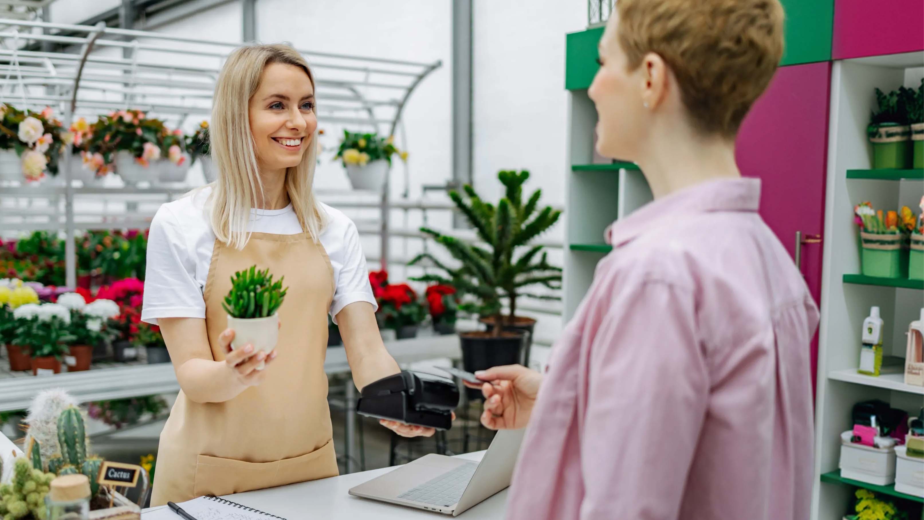 Verkäuferin übergibt einer Kundin im stationären Geschäft eine Pflanze und bietet digitale Bezahlung an.