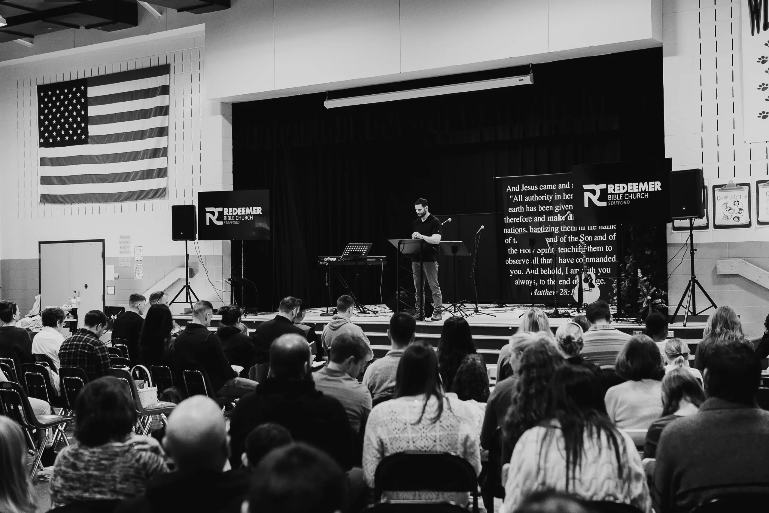 Congregation seated and listening to a speaker on stage at Redeemer Bible Church with U.S. flag and church signage in the background.