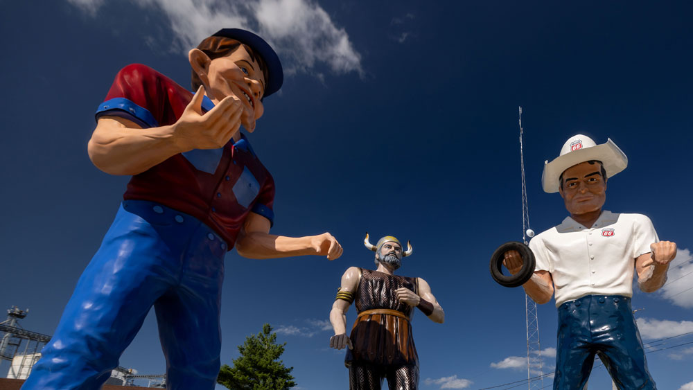 Three large vintage roadside statues against a blue sky: a man in a red and blue outfit, a Viking with a horned helmet, and a man in a white shirt and cowboy hat holding a tire.