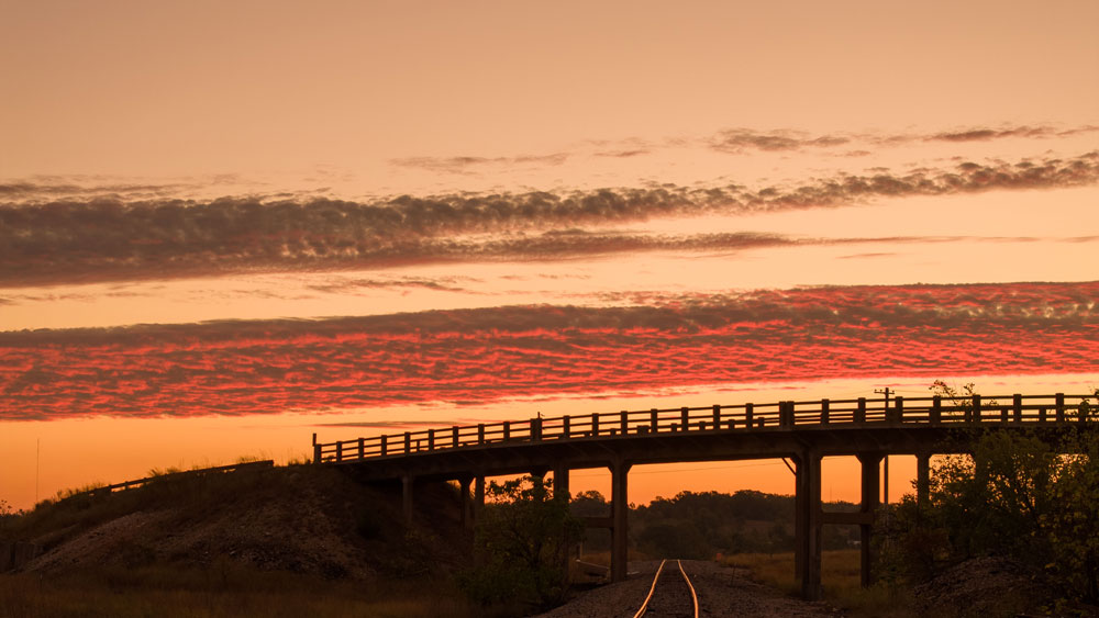 Railroad tracks running under a bridge at sunset with a colorful orange and red sky.