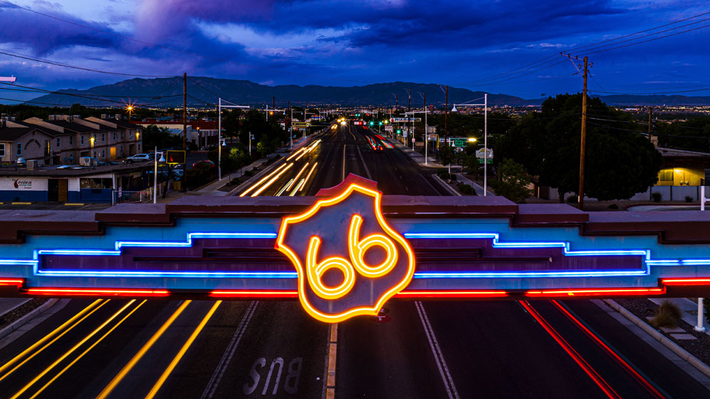 Neon-lit Route 66 sign over a busy highway at dusk with light trails from vehicles and mountains in the background.