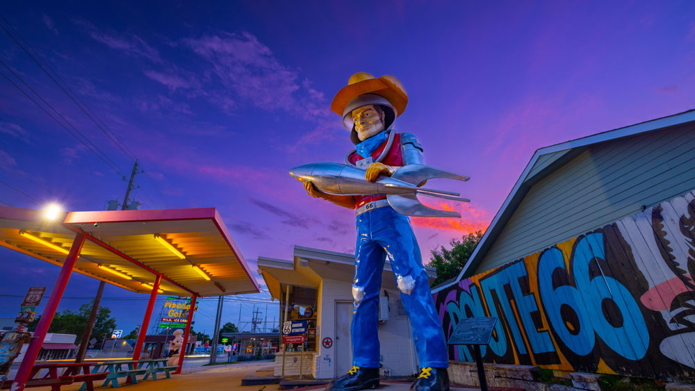 Large statue of a cowboy holding a silver rocket ship at dusk near a Route 66 mural and yellow-lit shelter.