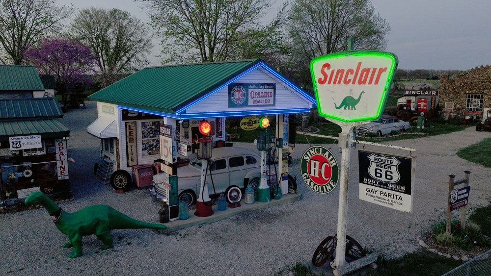 Vintage Sinclair gas station with neon-lit roof, classic pumps, a green dinosaur sculpture, and Route 66 signage on a gravel lot.