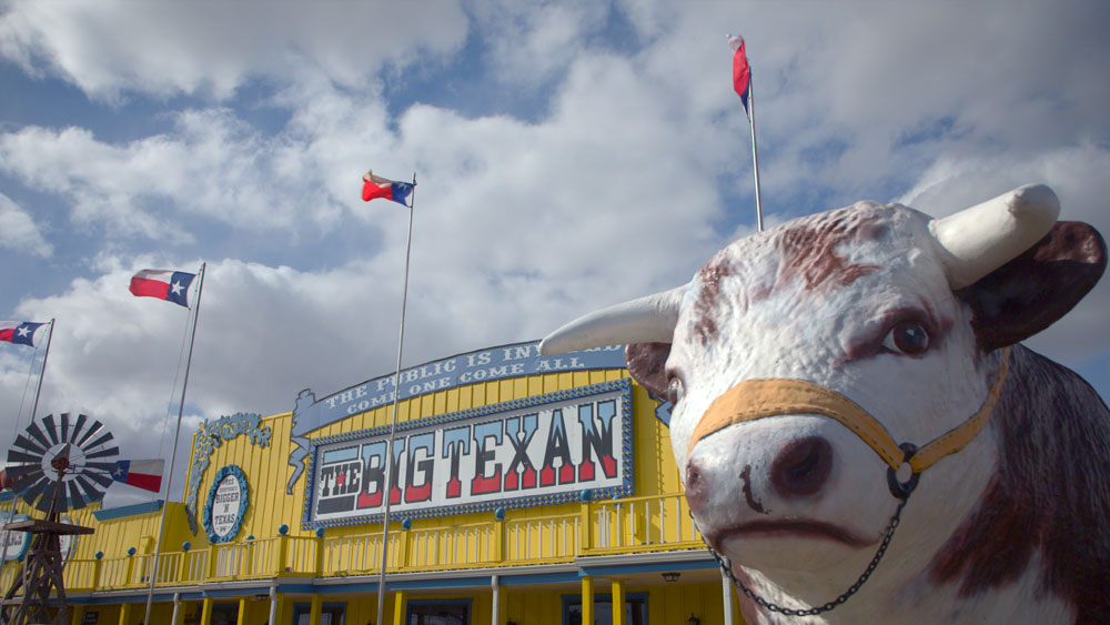 Facade of The Big Texan restaurant with Texas flags flying and a large bull statue in the foreground.