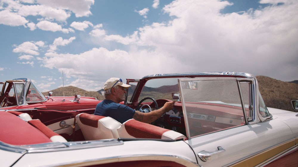 Older man wearing a beige cap and sunglasses driving a white vintage convertible car with red interior in a desert landscape under a partly cloudy sky.