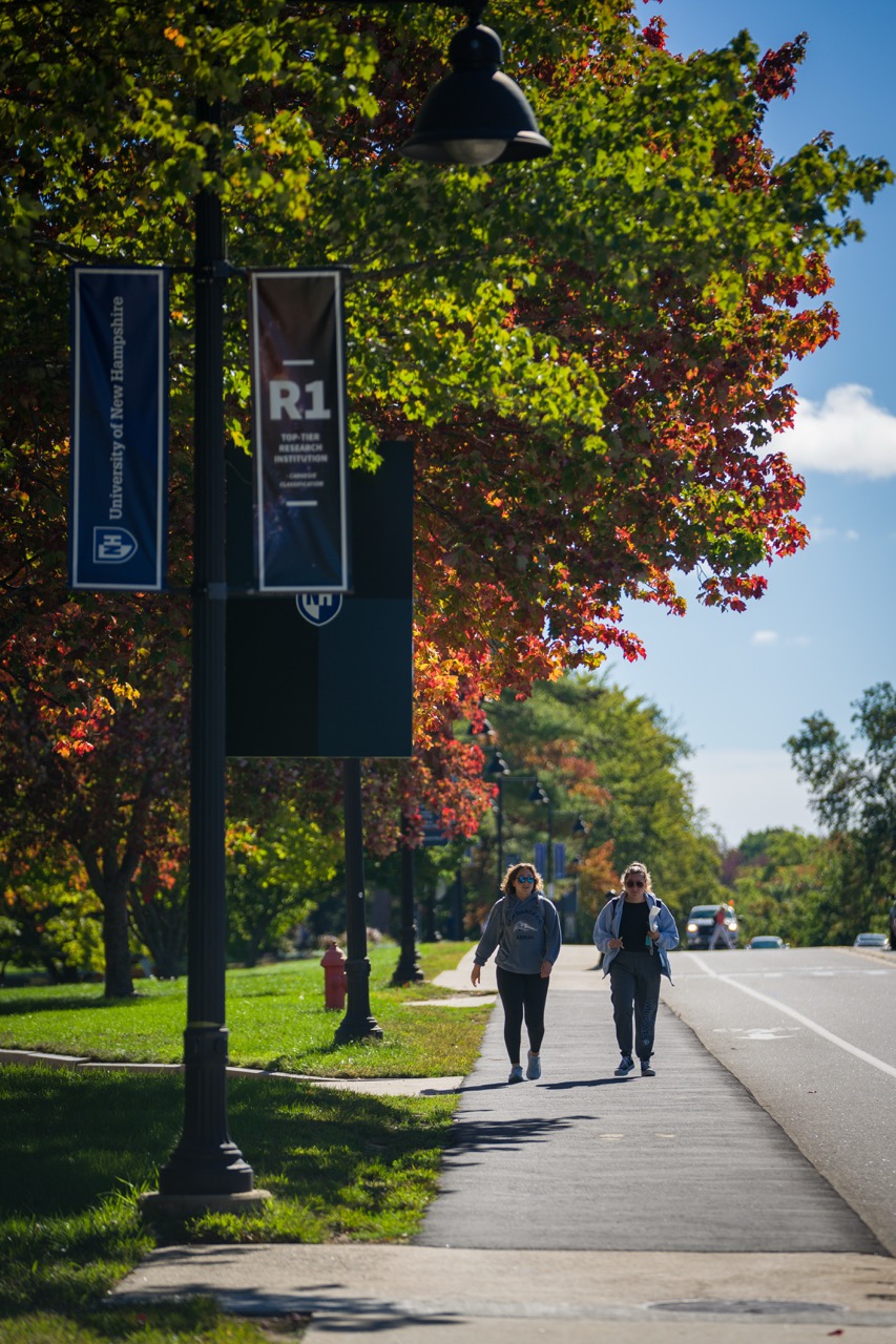 Two students walk along a tree-lined sidewalk on the University of New Hampshire campus, with fall foliage and campus banners visible under a sunny sky.