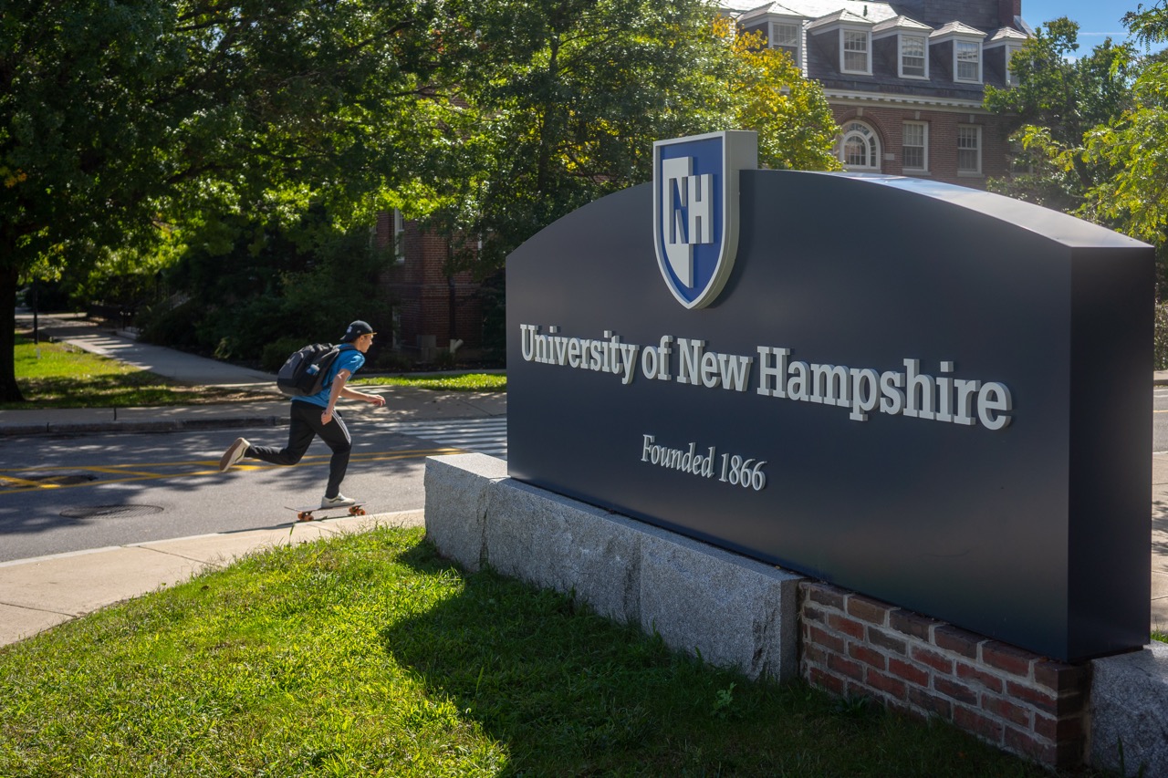 A male student on a skateboard passes by a large University of New Hampshire sign, which features the school's name and founding year, on a sunny day with campus buildings in the background.