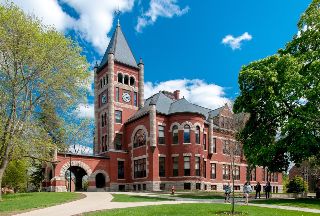 Thompson Hall, a red-brick building with a clock tower and arched entrance, on the University of New Hampshire campus surrounded by green trees and a bright blue sky with scattered clouds.