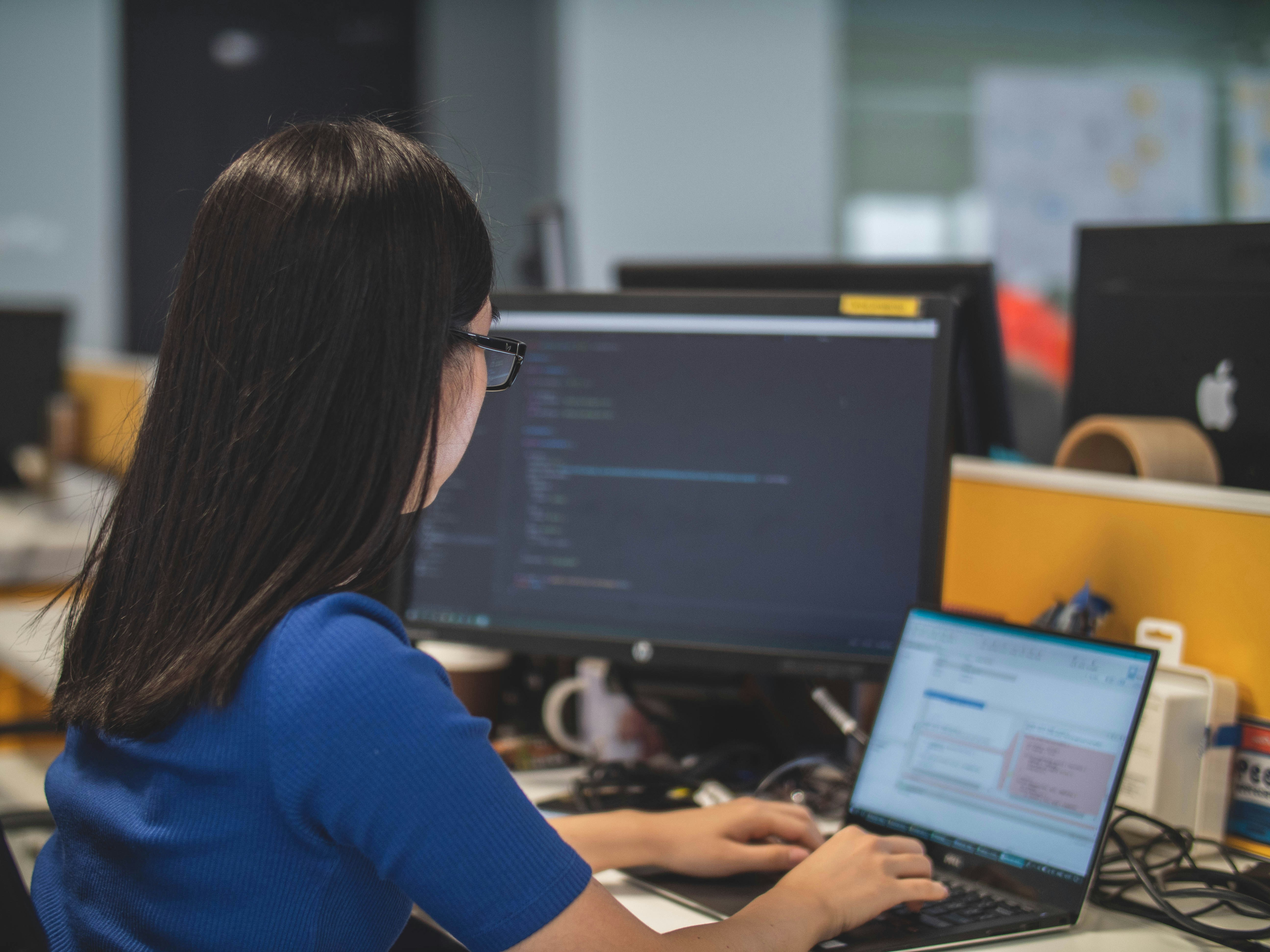 A female student in a blue t-shirt working at a desk with a laptop and large monitor displaying code.