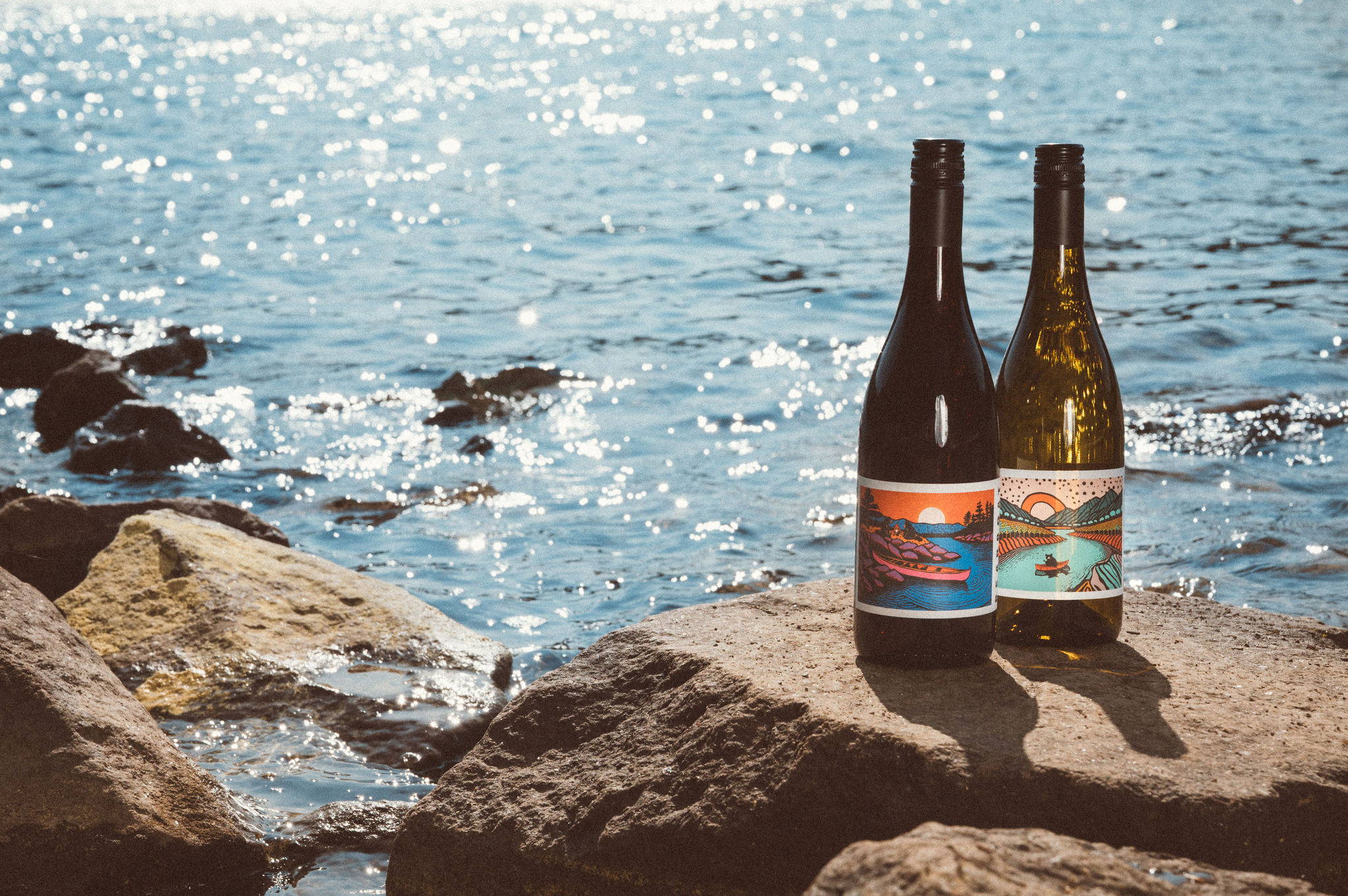 Bearboat Chardonnay and Pinot Noir bottles on sunlit rocks by the water