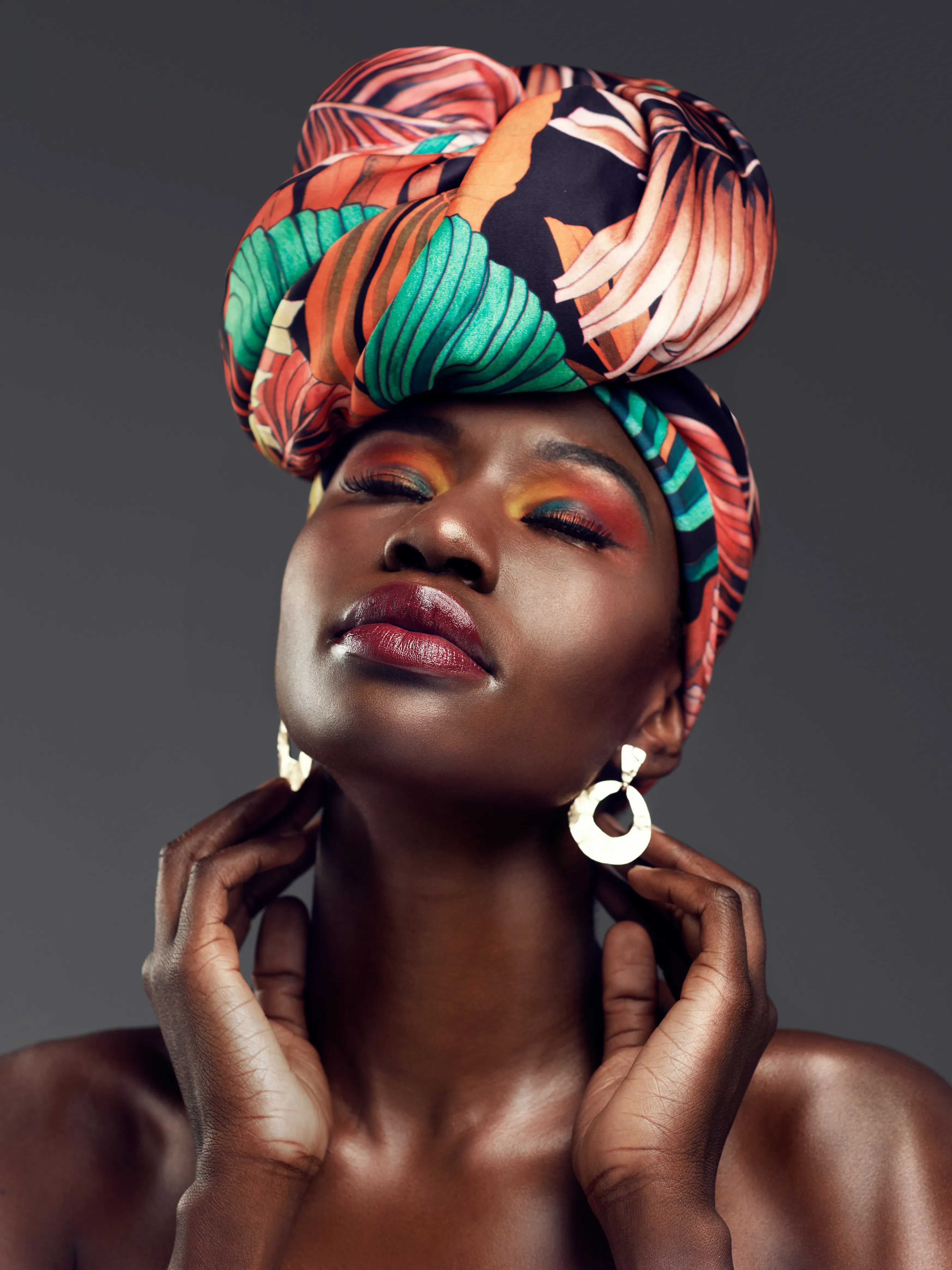 Woman in vibrant headwrap with colorful makeup poses elegantly against gray background