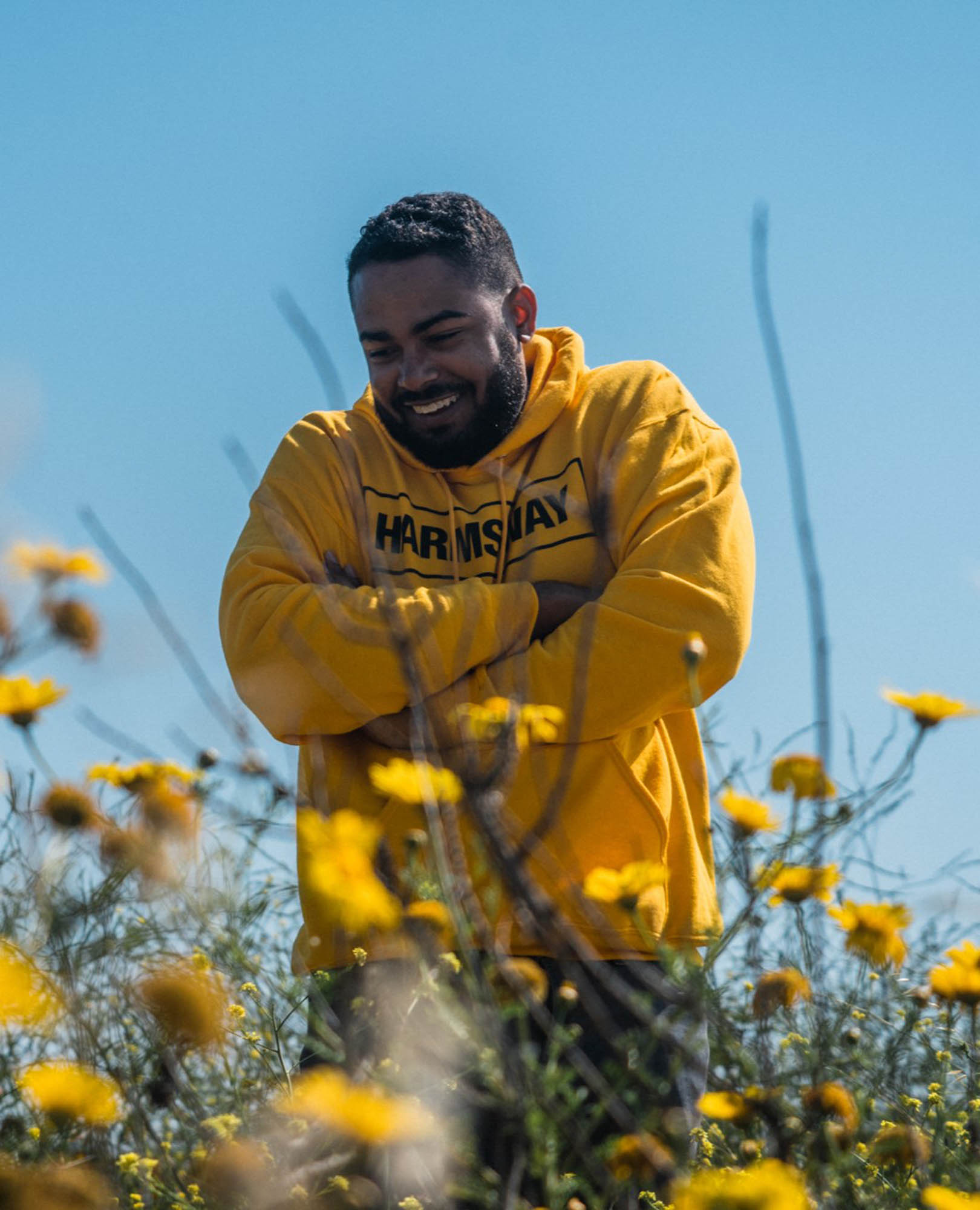 Comedian Curtis Cook smiling in a yellow hoodie standing among yellow flowers under a clear blue sky. Photography by Lex Voight