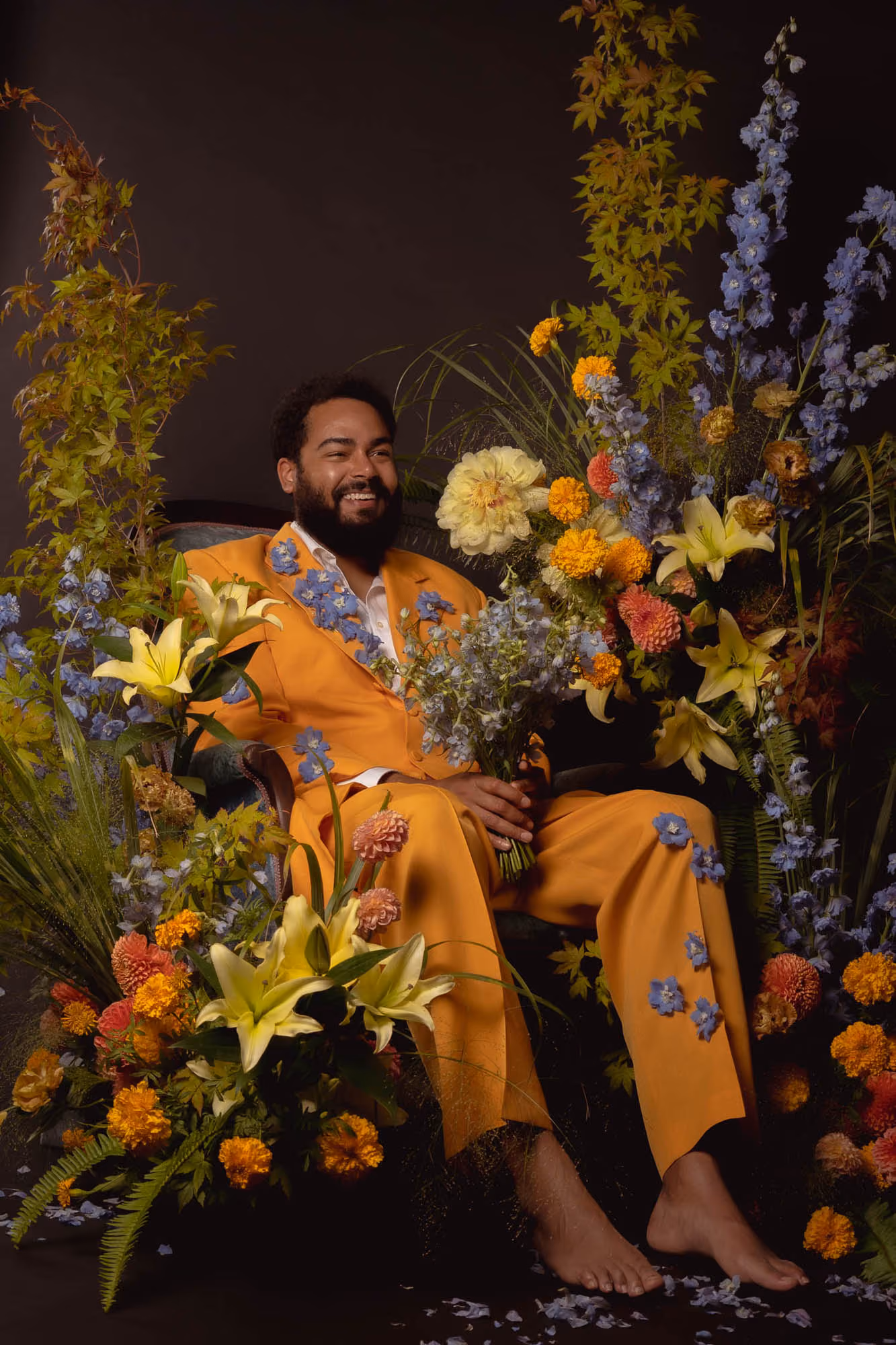 An image of comedian Curtis Cook wearing a yellow suit and sitting in a chair surrounded by flowers. Photography by Lex Voight and flowers by Nocturne in Bloom.