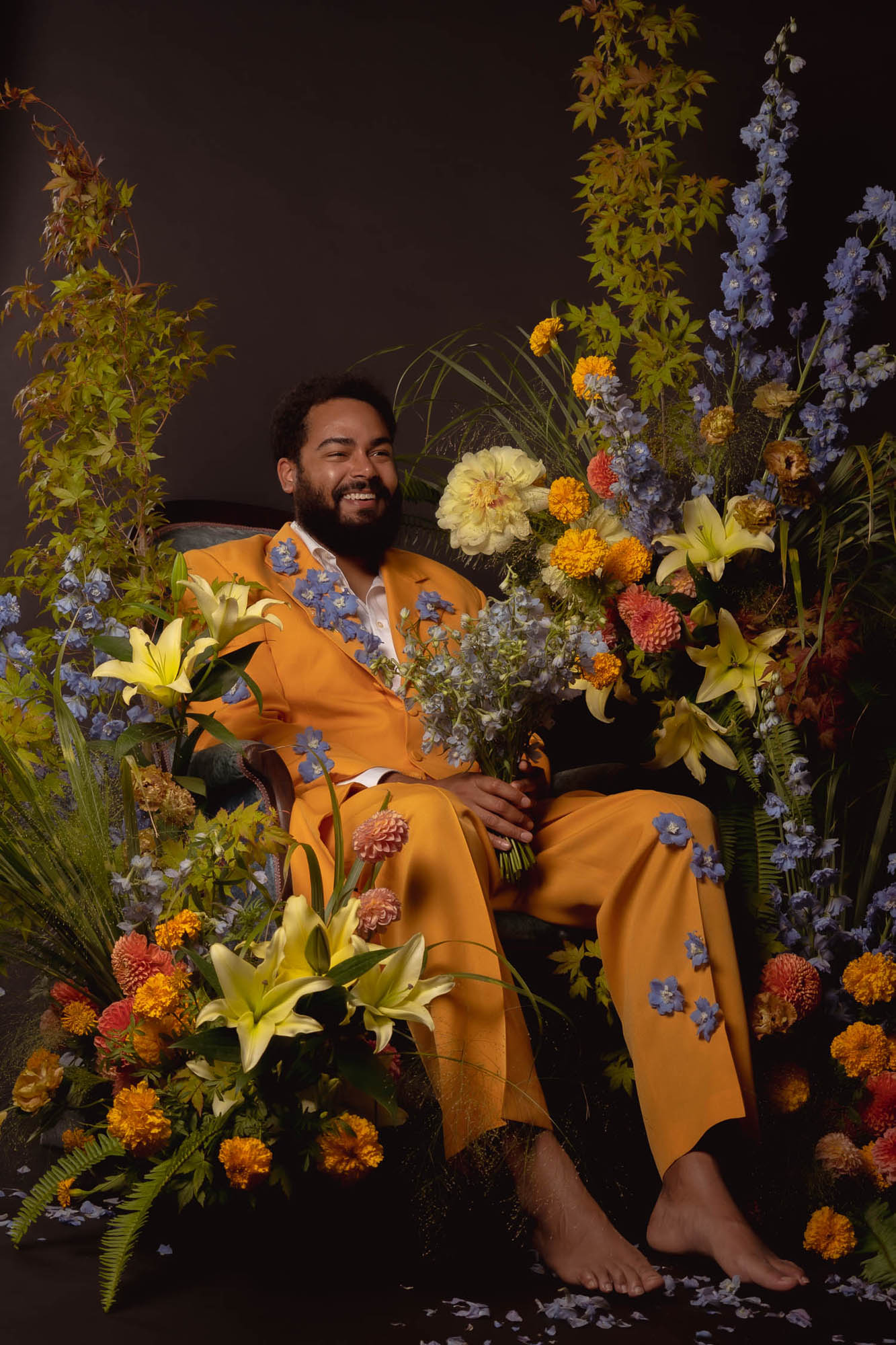 An image of comedian Curtis Cook wearing a yellow suit and sitting in a chair surrounded by flowers. Photography by Lex Voight and flowers by Nocturne in Bloom.