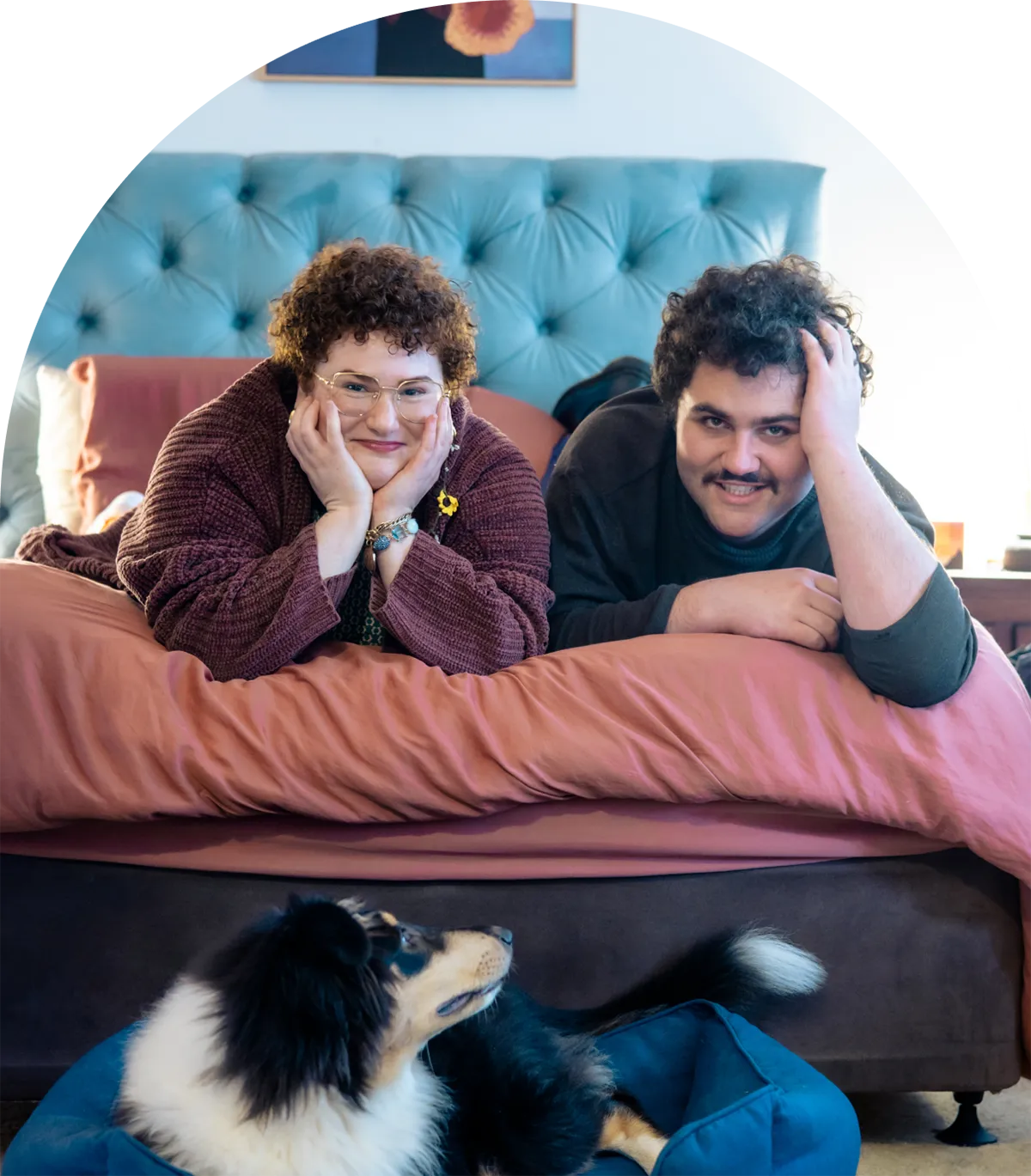 A photo of Emilia and their partner lying on their bed. Their dog can be seen lying on the floor in front of them